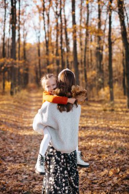 Açık gri bir kazak giyen ve parlak turuncu bir kazak giyen anne, renkli yapraklar ve uzun boylu yapraklarla çevrili şefkatli bir kucaklaşmayı paylaşırken sıcacık gülümsüyor. 