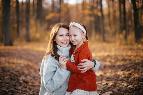 stock image Mother is hugging her daughter with braids in an autumn forest. The woman, dressed in a gray sweater and skirt, is hugging her little daughter, who is wearing a red sweater, in the autumn forest.