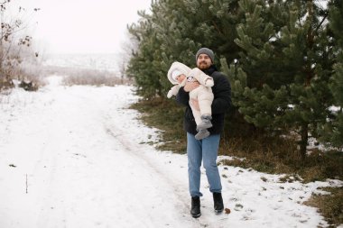 Sakallı adam beyaz bir tulum ve şapka giymiş, etrafı karla kaplı ve daima yeşil ağaçlarla çevrili gülümseyen bir bebek tutuyor. Babası ve küçük kızı arasında kışın neşeli bir an.