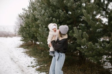 Kahverengi kısa kuzu derisi ceketli bir kadın, beyaz bir tulum ve şapka giymiş, karla kaplı zemin ve yemyeşil ağaçlarla çevrili bir bebeği kucağında tutuyor. Anne ve bebek arasında neşeli bir an. 