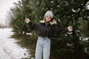 Happy woman is wearing a dark brown sheepskin jacket and a gray beanie while standing near evergreen trees in a snowy landscape. Enjoying the cold weather as she stretches her a arms out playfully.  clipart