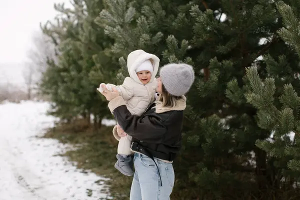 stock image A woman in a brown jacket, jeans, and a grey hat is spinning a baby in a white overall near evergreen trees in a snowy outdoor setting. Its a joyful winter scene of a mother and her little daughter.