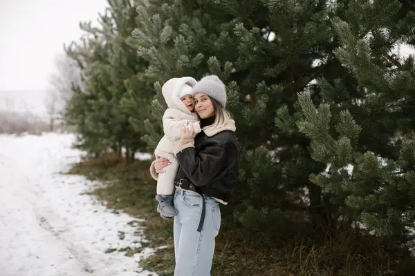 Kahverengi kısa kuzu derisi ceketli bir kadın, beyaz bir tulum ve şapka giymiş, karla kaplı zemin ve yemyeşil ağaçlarla çevrili bir bebeği kucağında tutuyor. Anne ve bebek arasında neşeli bir an. 