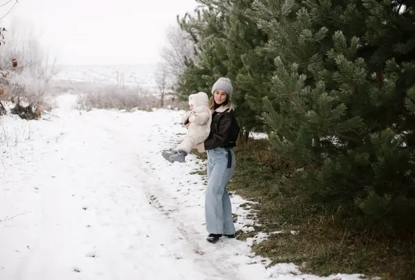 stock image A woman in a brown jacket, jeans, and a grey hat is spinning a baby in a white overall near evergreen trees in a snowy outdoor setting. Its a joyful winter scene of a mother and her little daughter. 