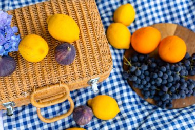 Mavi kareli masa örtüsüyle beyaz bir piknik sahnesi, narin bir ortanca, dokunmuş bir saman bavulu ve limon, portakal, incir ve taze üzümler içeren canlı meyveler.. 