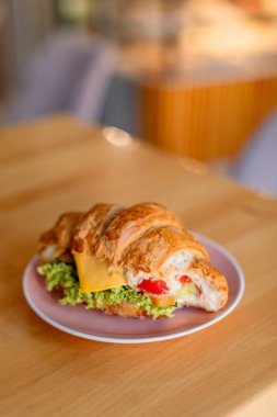 Pembe bir tabakta, ahşap bir masaya yerleştirilmiş Roissant sandviçi. Kruvasan marul, bir dilim peynir, mayonez ve parlak kırmızı biberle dolu. Renkli ve iştah açıcı bir görünüm yaratıyor.. 