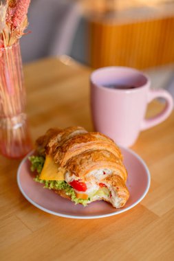 Peynir, marul ve sebzelerle dolu pembe bir tabakta servis edilen rossant sandviçi. Arkasında, sıcak ve bulanık bir arkaplanı olan yumuşak ve rahat bir atmosfer yaratan uyumlu pembe bir kupa var..