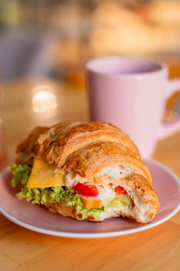 Peynir, marul ve sebzelerle dolu pembe bir tabakta servis edilen rossant sandviçi. Arkasında, sıcak ve bulanık bir arkaplanı olan yumuşak ve rahat bir atmosfer yaratan uyumlu pembe bir kupa var..