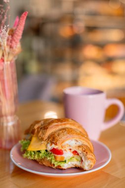 Peynir, marul ve sebzelerle dolu pembe bir tabakta servis edilen rossant sandviçi. Arkasında, sıcak ve bulanık bir arkaplanı olan yumuşak ve rahat bir atmosfer yaratan uyumlu pembe bir kupa var..