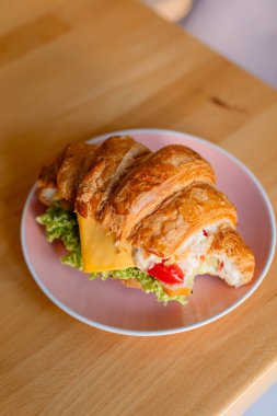 Pembe bir tabakta, ahşap bir masaya yerleştirilmiş Roissant sandviçi. Kruvasan marul, bir dilim peynir, mayonez ve parlak kırmızı biberle dolu. Renkli ve iştah açıcı bir görünüm yaratıyor.. 