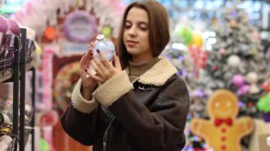 Kısa kahverengi koyun derisi ceket giyen uzun saçlı genç bir kadın bir mağazadan Noel süsleri seçiyor. Elinde mavi bir Noel süsü tutuyor..