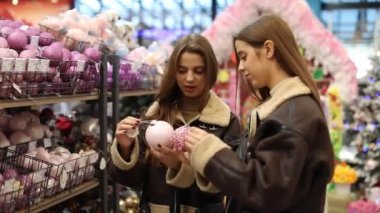 Kısa kahverengi püsküllü paltolar giyen ikiz kardeşler, bayram süslemeleriyle çevrili bir mağazadan Noel süsleri seçiyorlar. İki genç kadın Noel ağacı süslemelerini seçiyor.. 