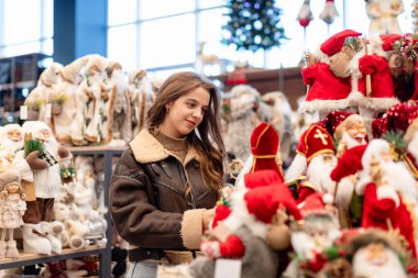 A young woman with long hair in a short brown sheepskin coat chooses Christmas decorations in a store. The woman holds a figure of St. Nicholas or Santa Claus in her hands. clipart