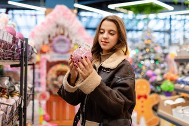 Kısa kahverengi koyun derisi ceket giyen uzun saçlı genç bir kadın bir mağazadan Noel süsleri seçiyor. Elinde pembe bir Noel süsü tutuyor..