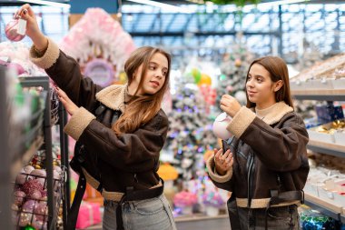 Kısa kahverengi püsküllü paltolar giyen ikiz kardeşler, bayram süslemeleriyle çevrili bir mağazadan Noel süsleri seçiyorlar. İki genç kadın Noel ağacı süslemelerini seçiyor.. 