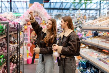 Kısa kahverengi püsküllü paltolar giyen ikiz kardeşler, bayram süslemeleriyle çevrili bir mağazadan Noel süsleri seçiyorlar. İki genç kadın Noel ağacı süslemelerini seçiyor.. 