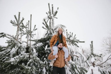 Neşeli bir çift, karlı bir kış gününün tadını çıkarıyor. Kadın, karla kaplı çam ağaçlarının arasında erkeğin omuzlarında şakacı bir şekilde oturuyor. Sahne, dingin, beyaz bir arkaplan karşısında sıcaklık ve neşe yakalıyor..