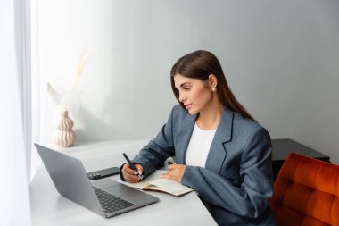 Masasında çalışan spor ceketli bir kadın dizüstü bilgisayarına bakarken elinde kalem tutuyor. Önünde bir defter açık duruyor ve içinde kurumuş bitkiler olan bir vazo boşluğu süsler. Boşluğu kopyala. 