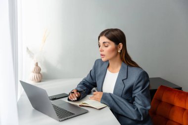 Masasında çalışan spor ceketli bir kadın dizüstü bilgisayarına bakarken elinde kalem tutuyor. Önünde bir defter açık duruyor ve içinde kurumuş bitkiler olan bir vazo boşluğu süsler. Boşluğu kopyala. 