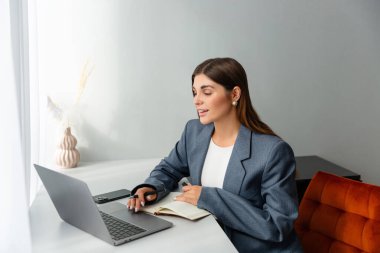 Masasında çalışan spor ceketli bir kadın dizüstü bilgisayarına bakarken elinde kalem tutuyor. Önünde bir defter açık duruyor ve içinde kurumuş bitkiler olan bir vazo boşluğu süsler. Boşluğu kopyala. 