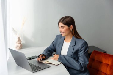 Masasında çalışan spor ceketli bir kadın dizüstü bilgisayarına bakarken elinde kalem tutuyor. Önünde bir defter açık duruyor ve içinde kurumuş bitkiler olan bir vazo boşluğu süsler. Boşluğu kopyala. 