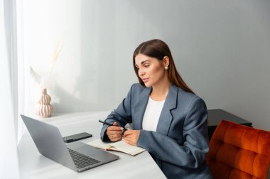 Masasında çalışan spor ceketli bir kadın dizüstü bilgisayarına bakarken elinde kalem tutuyor. Önünde bir defter açık duruyor ve içinde kurumuş bitkiler olan bir vazo boşluğu süsler. Boşluğu kopyala. 