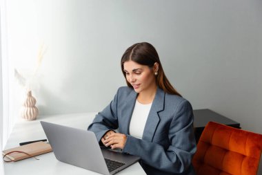 Gri ceketli bir kadın çevrimiçi konferans sırasında dizüstü bilgisayarına bakıyor ve gülümsüyor. İş yeri iyi ışıklandırılmış ve bir vazo kurumuş bitki estetik cazibe katıyor. Boşluğu kopyala. 