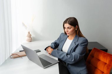 Beyaz bir masada dizüstü bilgisayar üzerinde çalışan profesyonel spor ceketli bir kadın. Kurutulmuş bitkilerle dekoratif bir vazo yakına yerleştirilir ve çalışma alanına samimi bir dokunuş eklenir.