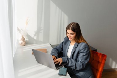 Woman in a professional blazer working on a laptop at a white desk. A decorative vase with dried plants is placed nearby, adding a cozy touch to the workspace. clipart