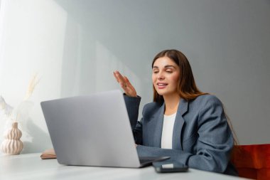 Gri ceketli bir kadın çevrimiçi konferans sırasında dizüstü bilgisayarına bakıyor ve gülümsüyor. İş yeri iyi ışıklandırılmış ve bir vazo kurumuş bitki estetik cazibe katıyor. Boşluğu kopyala. 