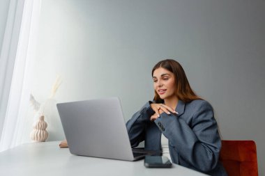 Gri ceketli bir kadın çevrimiçi konferans sırasında dizüstü bilgisayarına bakıyor ve gülümsüyor. İş yeri iyi ışıklandırılmış ve bir vazo kurumuş bitki estetik cazibe katıyor. Boşluğu kopyala. 