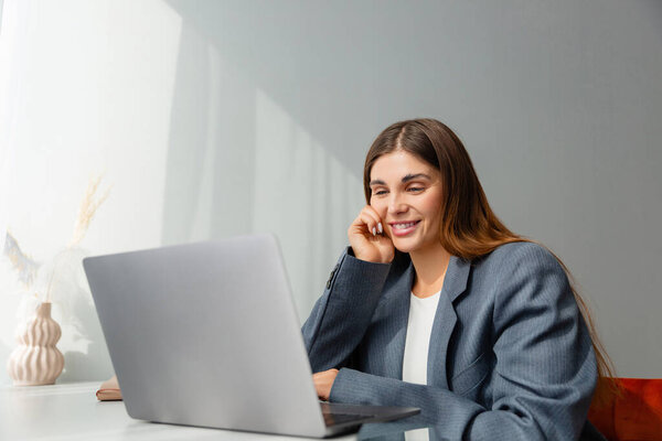 Женщина в сером пиджаке смотрит на свой ноутбук и улыбается во время онлайн-конференции. Рабочее место хорошо освещено, а ваза из сушеных растений добавляет эстетической привлекательности. Копирование пространства. 