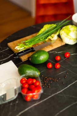 Ahşap kesme tahtasındaki yeşil soğanları mutfak bıçağıyla doğrayan ellerin yakın çekimi. Arka planda marul ve avokado gibi taze malzemeler sağlıklı bir pişirme atmosferi yaratır..