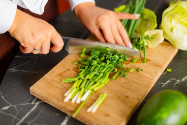 Close-up of hands slicing green onions on a wooden cutting board with a kitchen knife.Fresh ingredients like lettuce and avocado in the background create a healthy cooking atmosphere. clipart