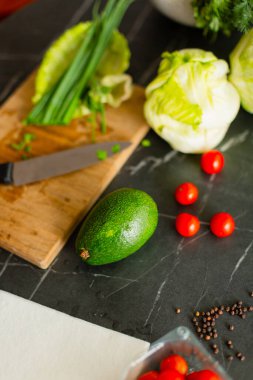 Ahşap kesme tahtasındaki yeşil soğanları mutfak bıçağıyla doğrayan ellerin yakın çekimi. Arka planda marul ve avokado gibi taze malzemeler sağlıklı bir pişirme atmosferi yaratır..