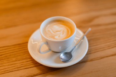 Beyaz seramik bardakta servis edilen ve gümüş kaşıkla eşleşen bir fincan içinde servis edilen narin latteyle klasik bir kapuçino. Sıcak ahşap masa sıcak bir kahve dükkanı ortamı ekler..