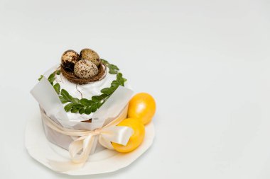 Beyaz kremayla süslenmiş tek bir Paskalya pastası, altın yumurtalarla dolu küçük bir yuva, taze yeşil yapraklar, ve zarif bir şenlik görüntüsü veren saten bir kurdele..