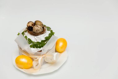 Beyaz kremayla süslenmiş tek bir Paskalya pastası, altın yumurtalarla dolu küçük bir yuva, taze yeşil yapraklar, ve zarif bir şenlik görüntüsü veren saten bir kurdele..
