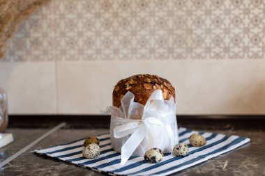 Geleneksel Paskalya ekmeği (kulich) üzerinde çikolata taneleri, saten kurdeleyle beyaz parşömen kağıdına sarılı vazo ve benekli bıldırcın yumurtalarıyla birlikte mavi çizgili bir havluya kuruludur..