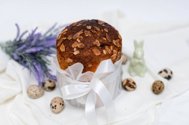 Geleneksel Paskalya ekmeği (kulich) üzerine beyaz parşömen kağıdına sarılı beyaz parşömen kurdelesi, yanında bıldırcın yumurtası ve mor lavanta filizi serpiştirilmiş açık kumaş arka plan..