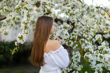 Beyaz bluzlu, uzun kahverengi saçlı genç bir kadın sırtı kameraya dönük duruyor çiçek açan kiraz ağacının altında nazikçe çiçeklere dokunuyor..