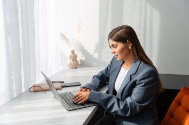 Gri ceketli genç bir kadın modern bir masada dizüstü bilgisayarı üzerinde çalışıyor. Görüntü üretkenliği, odaklanmayı ve uzak çalışmayı iletir.
