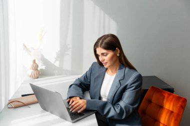 Gri ceketli genç bir kadın modern bir masada dizüstü bilgisayarı üzerinde çalışıyor. Görüntü üretkenliği, odaklanmayı ve uzak çalışmayı iletir.