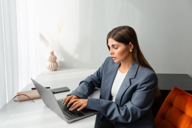 Gri ceketli genç bir kadın modern bir masada dizüstü bilgisayarı üzerinde çalışıyor. Görüntü üretkenliği, odaklanmayı ve uzak çalışmayı iletir.