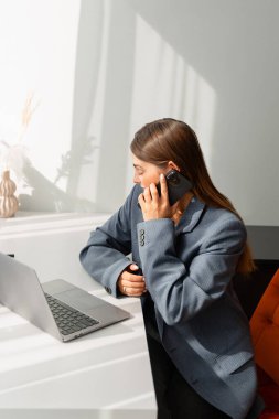 Gri ceketli genç bir kadın modern bir masada oturmuş, akıllı telefonuyla konuşuyor. Yanında bir dizüstü bilgisayar açık. İmaj iş iletişimi ve profesyonelliği aktarıyor.