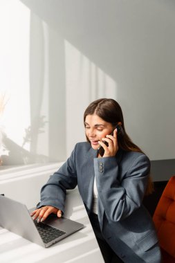 Gri ceketli genç bir kadın modern bir masada oturmuş, akıllı telefonuyla konuşuyor. Yanında bir dizüstü bilgisayar açık. İmaj iş iletişimi ve profesyonelliği aktarıyor.