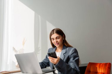 Gri ceketli genç bir kadın modern bir masada oturuyor, akıllı telefonunu kullanıyor. Yanında bir dizüstü bilgisayar açık. İmaj iş, iletişim ve üretkenlik taşır.