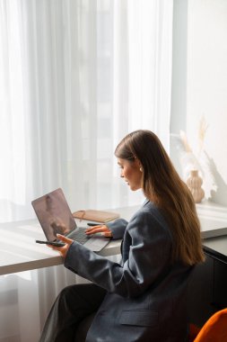 Gri ceketli genç bir kadın parlak, modern bir masada dizüstü bilgisayarda çalışıyor. Sahne üretkenlik, uzak çalışma ve profesyonelliği aktarıyor.
