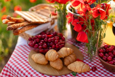 Taze kruvasan, kiraz, Grissini ve gelincik çiçekleri yaz otlağında kırmızı bir gingham masa örtüsüyle piknik masasında..