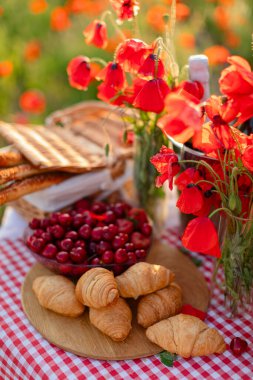 Taze kruvasan, kiraz, Grissini ve gelincik çiçekleri yaz otlağında kırmızı bir gingham masa örtüsüyle piknik masasında..